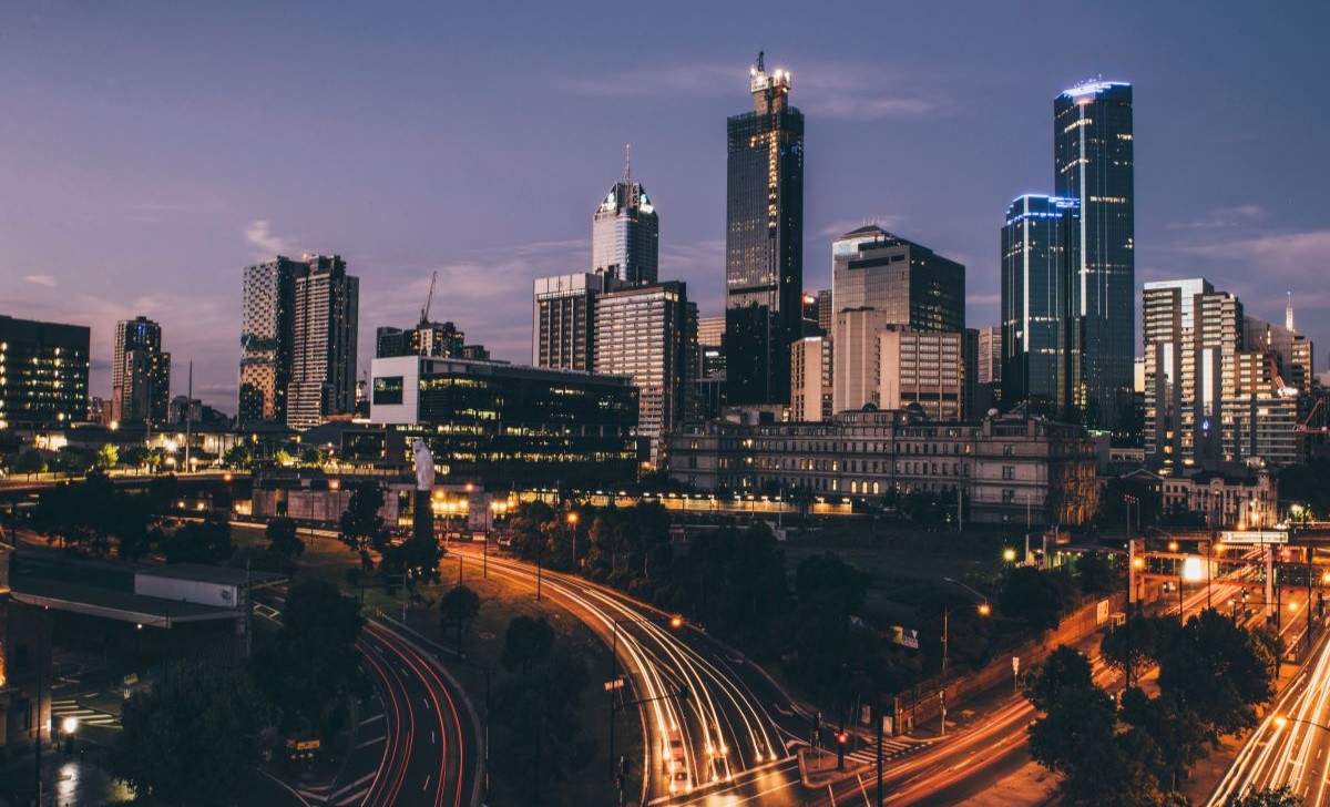 A nighttime cityscape with a variety of illuminated skyscrapers and buildings. In the foreground, there are roads with light trails, indicating moving traffic.